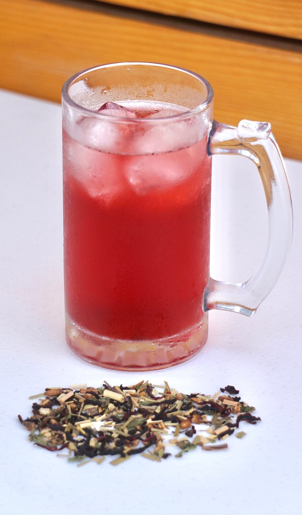 Sunny Hills Tea Ruby Glow - organic hibiscus, organic lemongrass, organic peppermint, dandelion leaf, roasted dandelion root. The tea brewed in this image is iced tea. Nice and cold for a hot summer day. And there is a hand full of the tea in loose leaf form on the table.